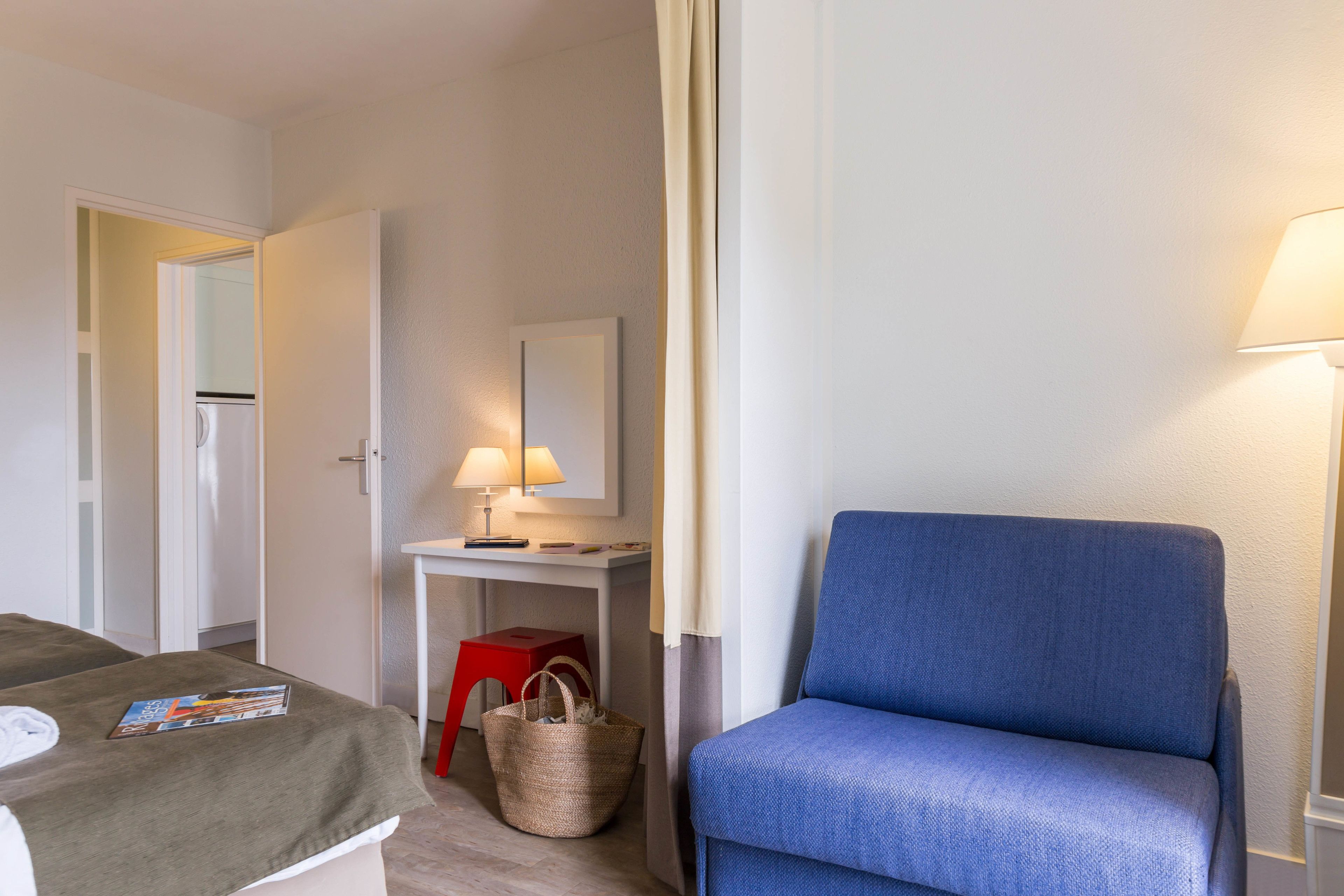 Una habitación de hotel con una cama, un escritorio con lámpara, una silla azul y un taburete rojo.