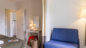 Una habitación de hotel con una cama, un escritorio con lámpara, una silla azul y un taburete rojo.