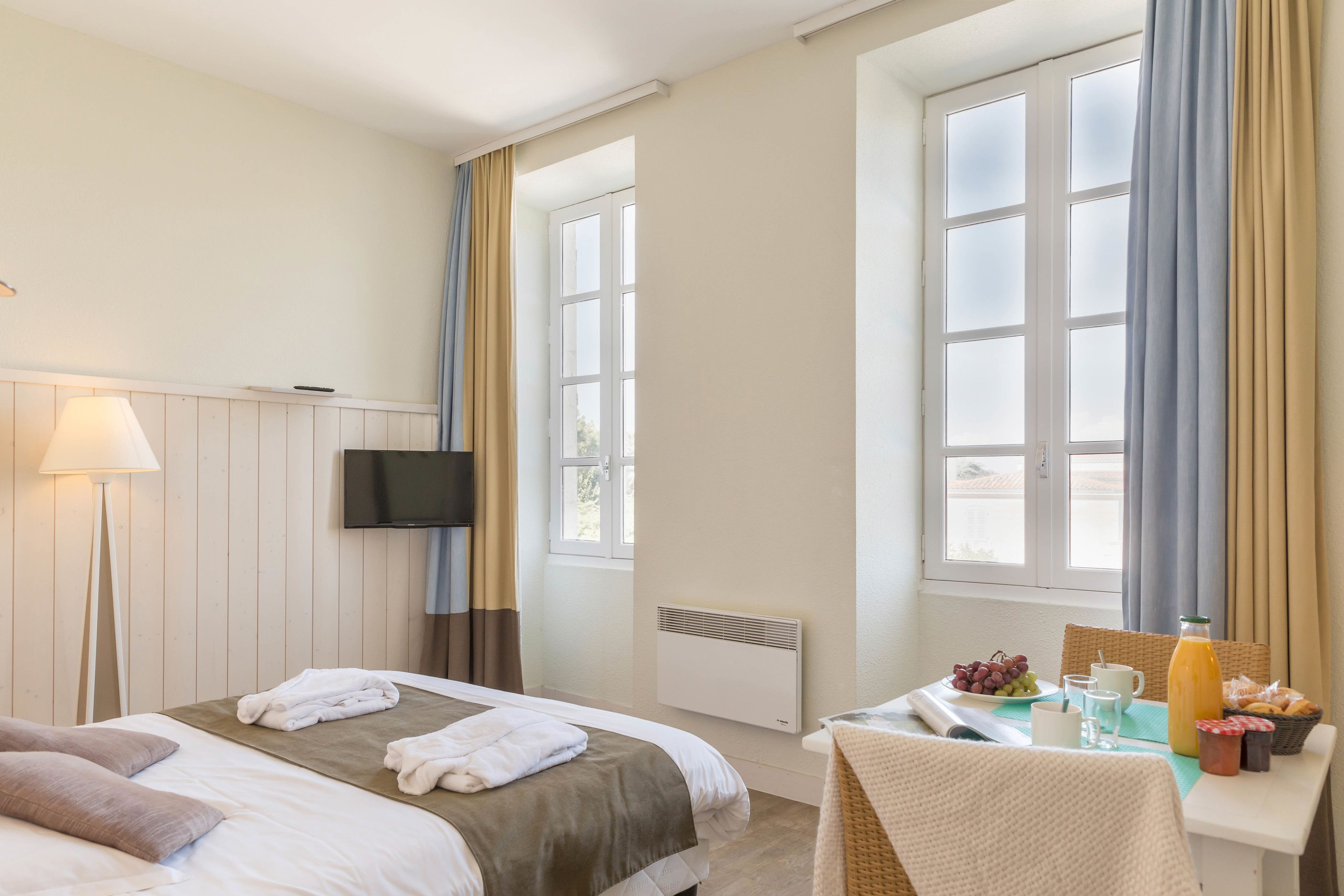A hotel room with a bed, a TV, a table with breakfast items, and a window with curtains.