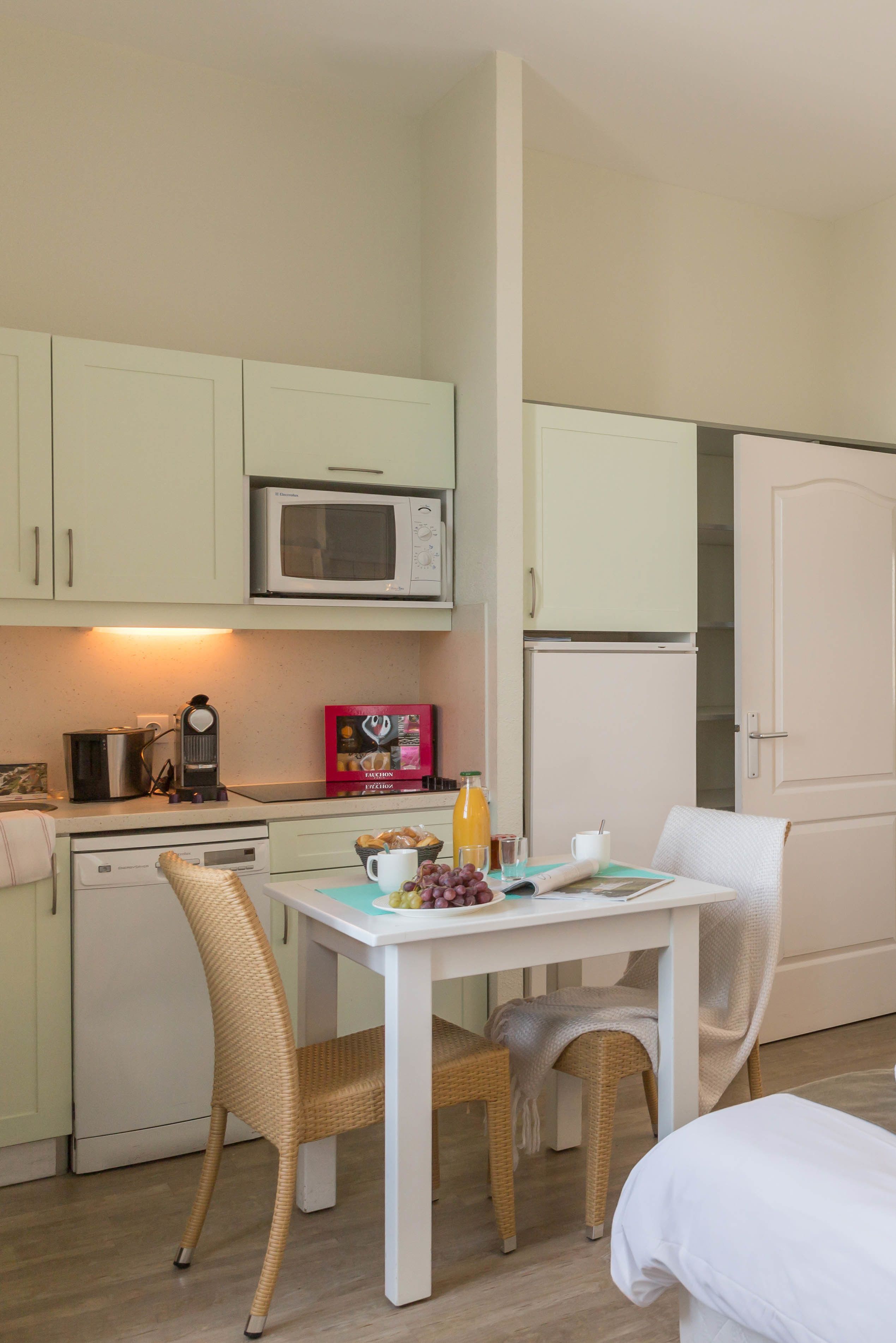 Cocina pequeña equipada con microondas, tostadora y refrigerador. Se aprecia una mesa de comedor con sillas de mimbre y mantel blanco.