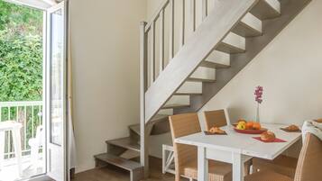 A modern interior with a staircase, a dining area with wicker chairs, and a table set with fruits and pastries.