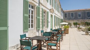 An outdoor café with wooden tables and green chairs, set against a white building with green shutters. - Résidence Le Palais des Gouverneurs Pierre & Vacances (Saint-Martin-de-Re)