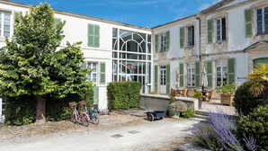 A courtyard with a white building, green shutters, a tree, bicycles, a wheelbarrow, and outdoor seating. - Résidence Le Palais des Gouverneurs Pierre & Vacances (Saint-Martin-de-Re)