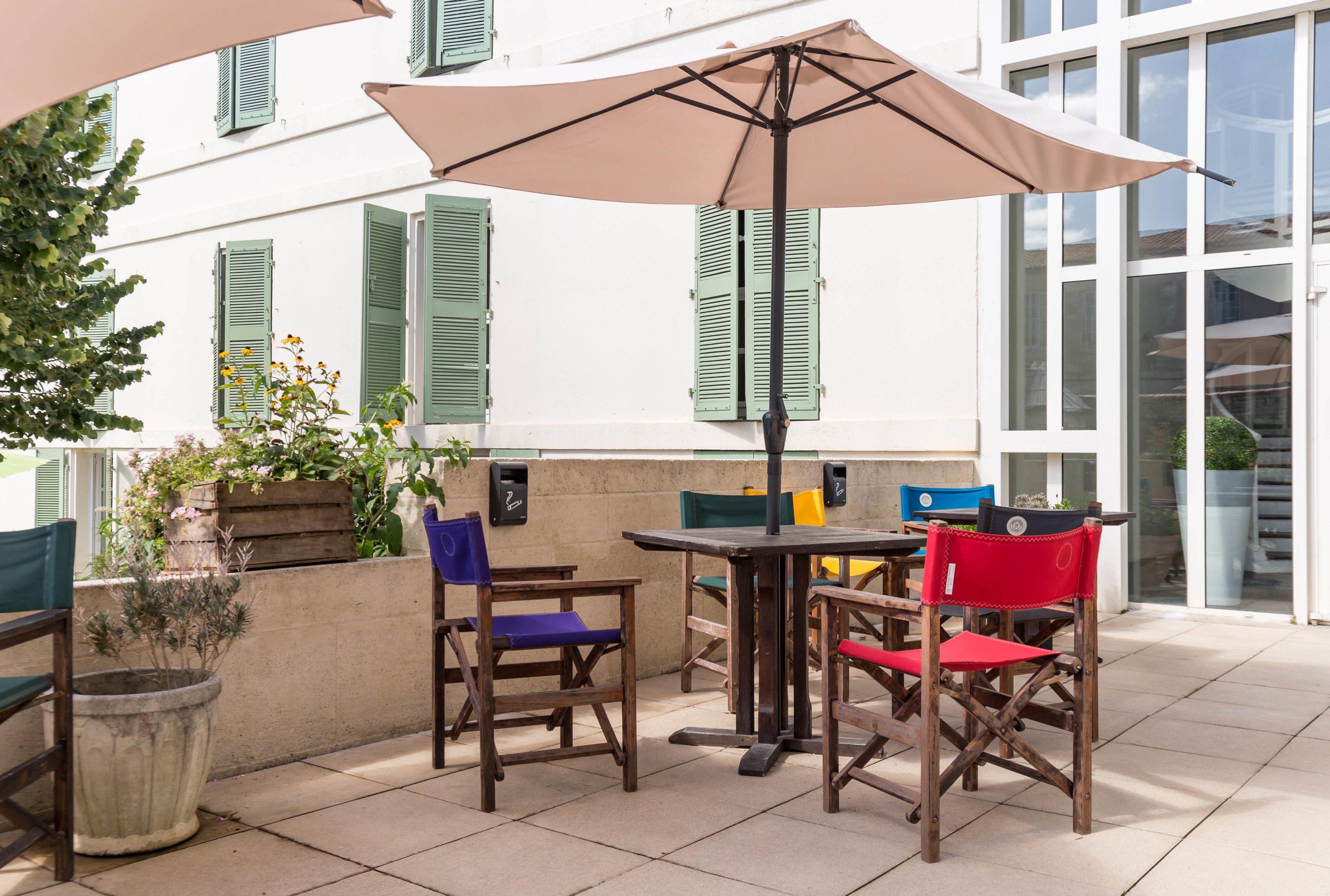 Een terras buiten met een tafel, stoelen, een grote parasol en een gebouw met groene luiken op de achtergrond.