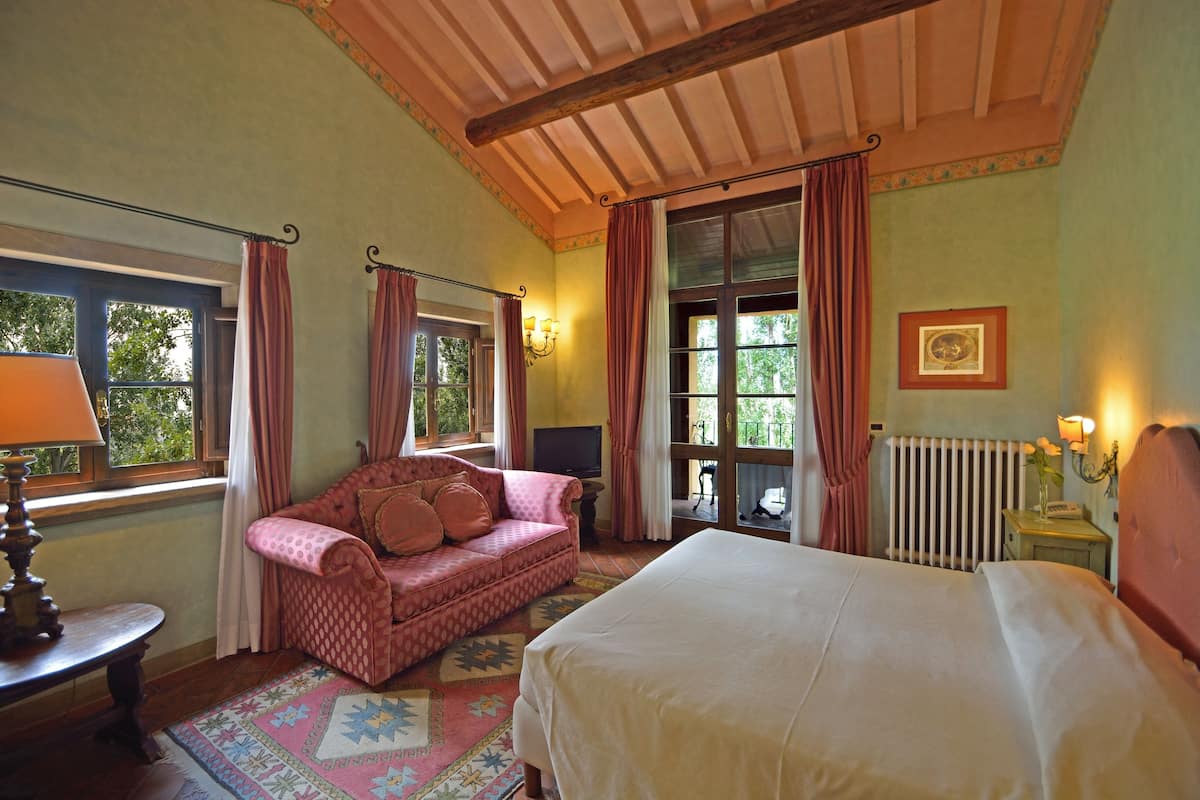 A bedroom with a bed, a sofa, a window with curtains, a radiator, and a wall-mounted lamp.
