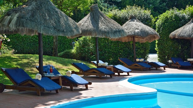 Área de piscina com espreguiçadeiras e guarda-sóis de palha, cercada por vegetação.