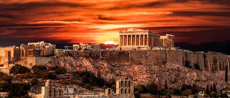 Un paisaje urbano histórico con ruinas antiguas y un edificio clásico destacado en una colina, bajo un cielo de atardecer espectacular.