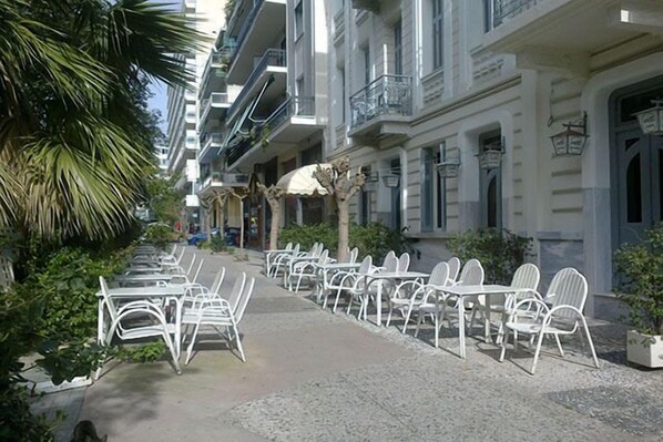 A row of white metal chairs and tables on a paved sidewalk, flanked by buildings with balconies and windows.