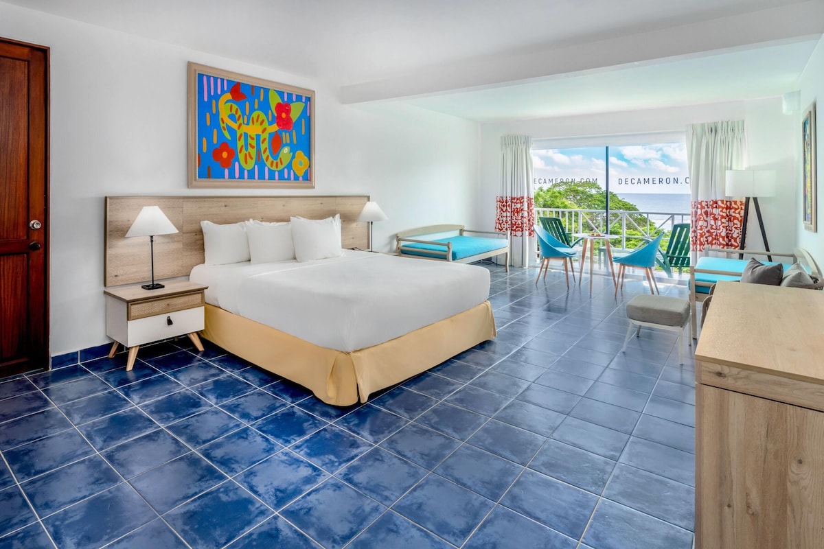 A hotel room with a large bed, a desk with chairs, a balcony with a view, and a colorful painting on the wall.