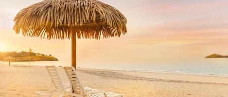 A beach scene with two white lounge chairs and a thatched umbrella.