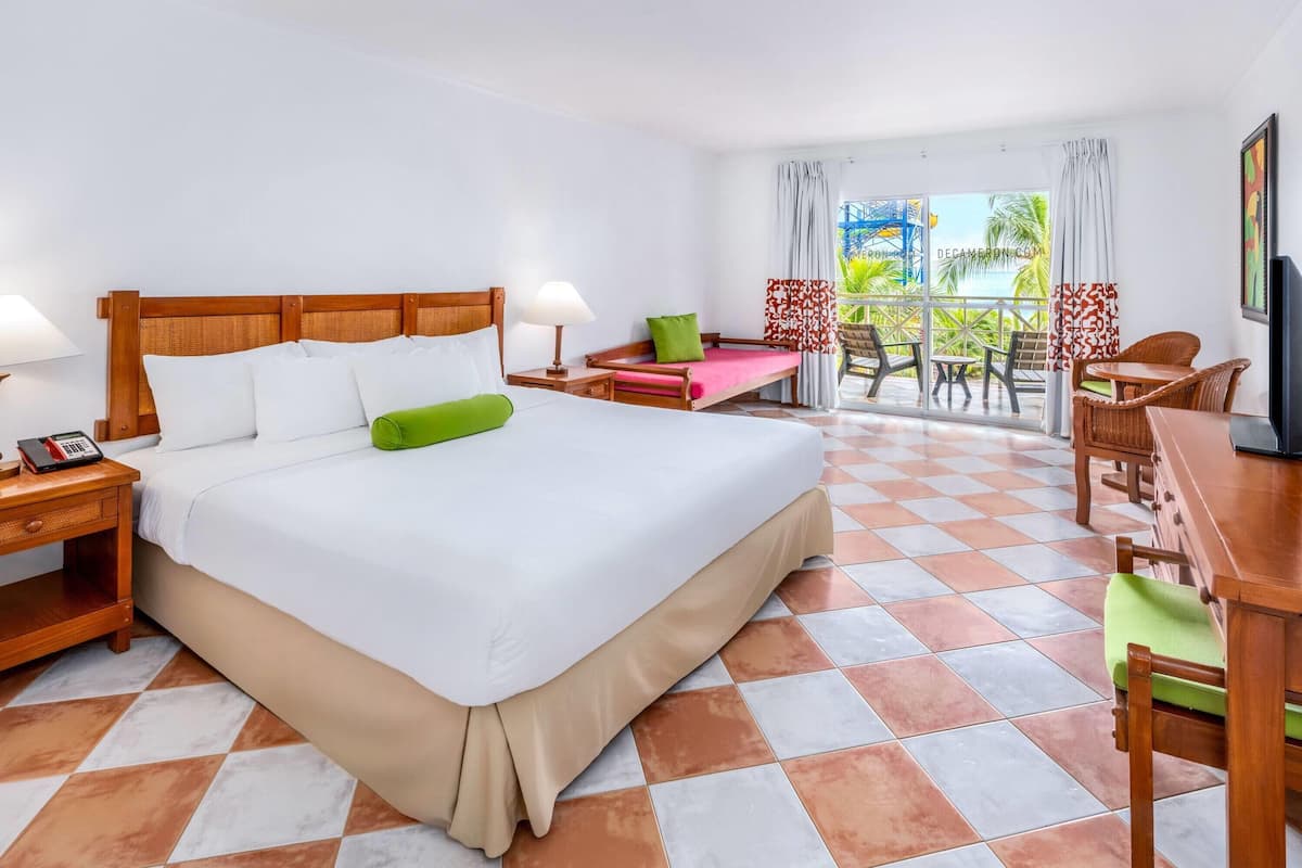 Een hotelkamer met een groot bed, een bureau, een stoel en een balkon met uitzicht op palmbomen en een blauwe constructie.
