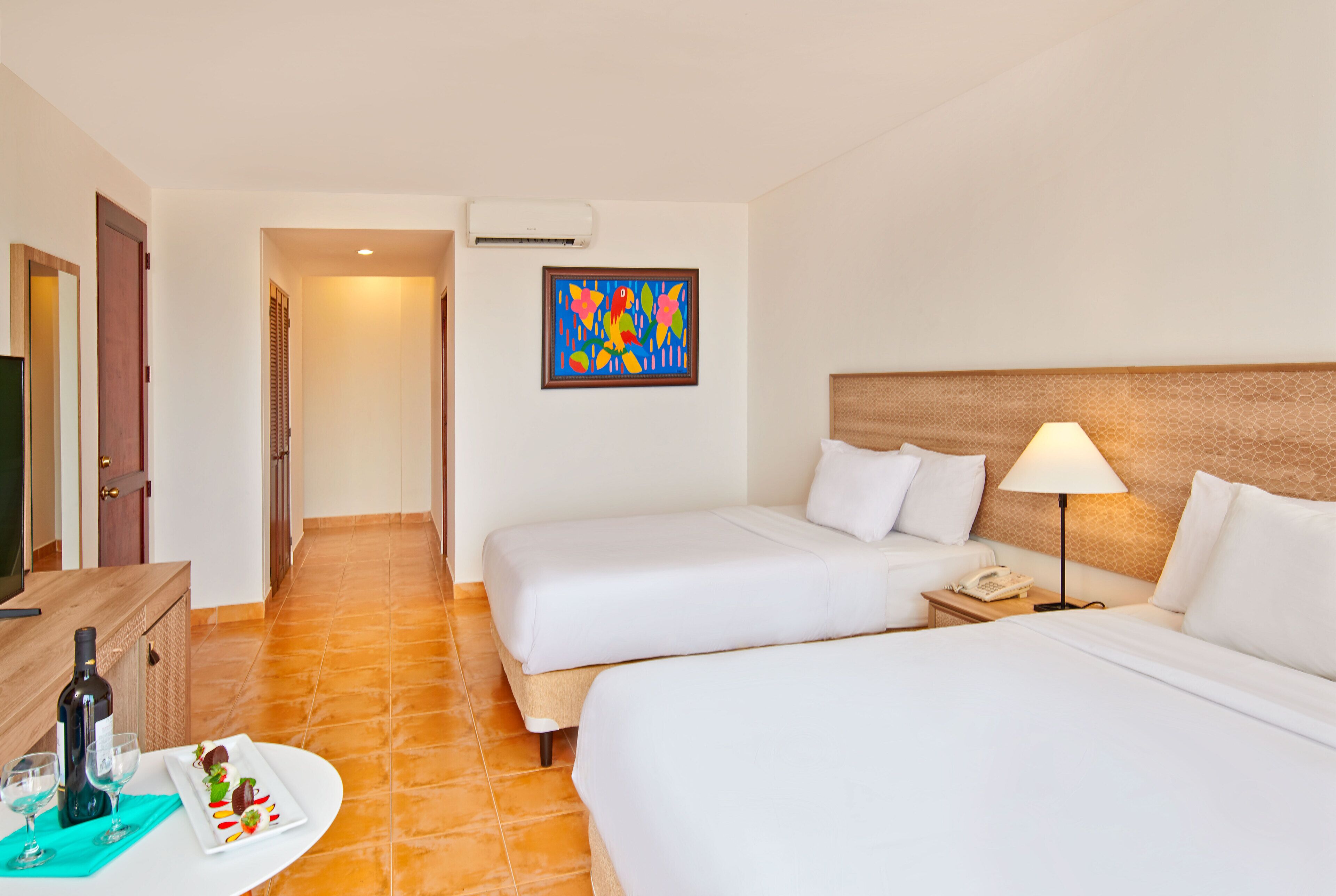 A hotel room with two beds, a desk with a bottle of wine, a plate of food, and a colorful painting on the wall.