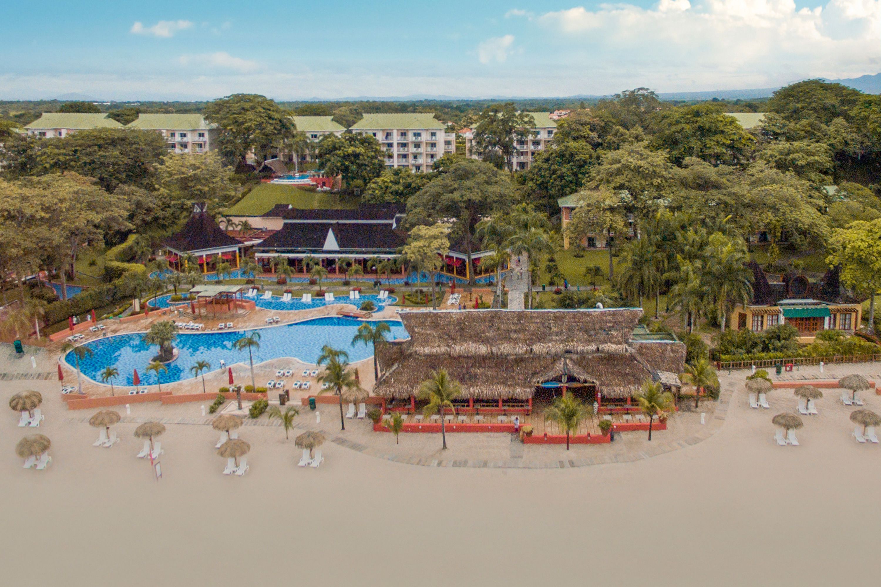 Un resort con piscina central, varias edificaciones y una playa con sombrillas y sillas reclinables.
