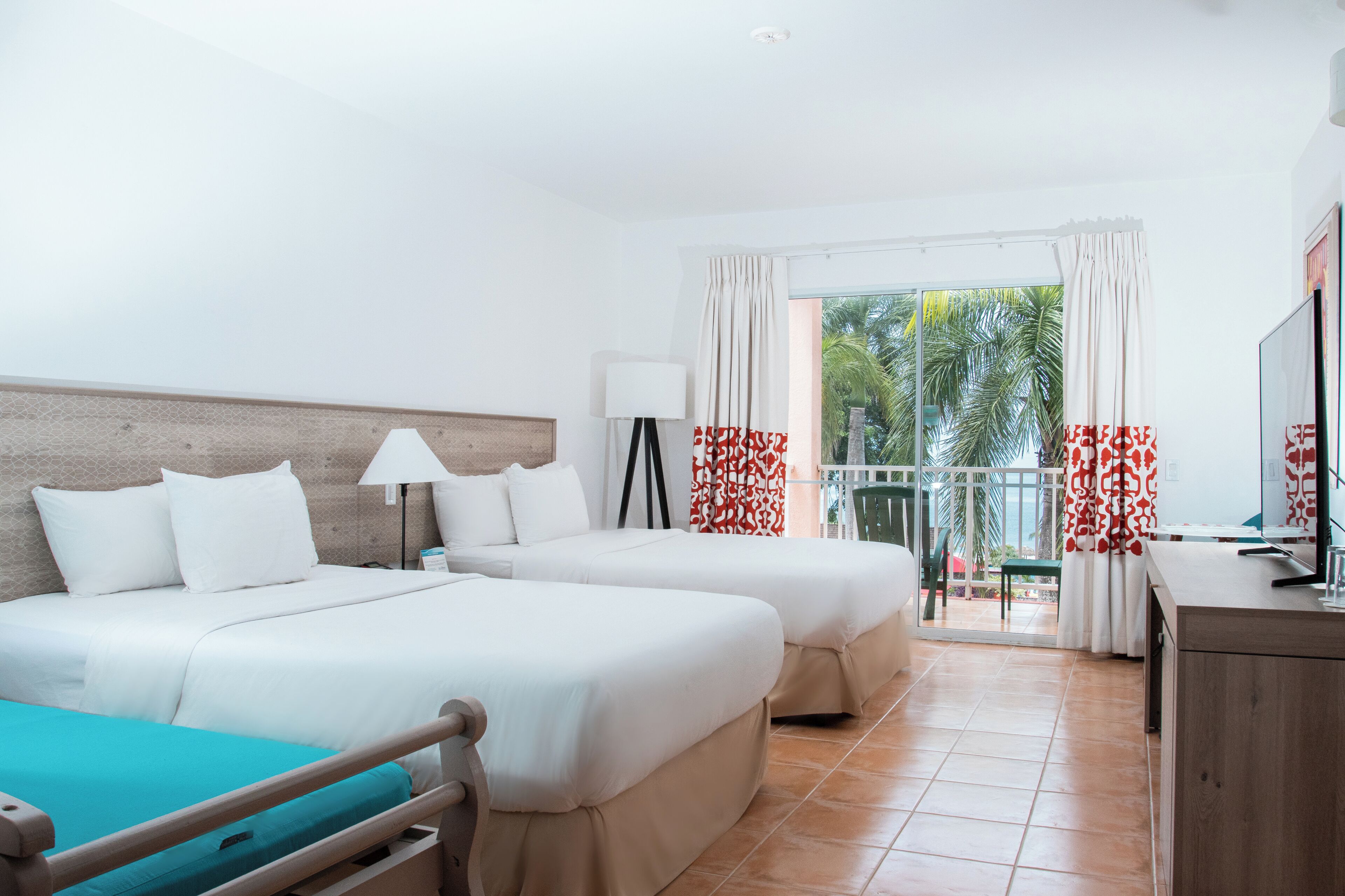 A hotel room with two beds, a desk, a TV, and a balcony with curtains.