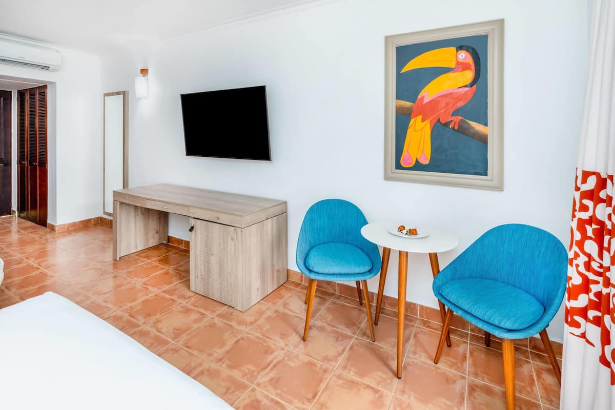 Een hotelkamer met een houten bureau, twee blauwe stoelen, een flatscreen televisie en een kleurrijk schilderij van een toekan op de muur.