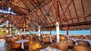 Un espacio de comedor al aire libre con sillas y mesas de mimbre, techo de paja y vista al océano.