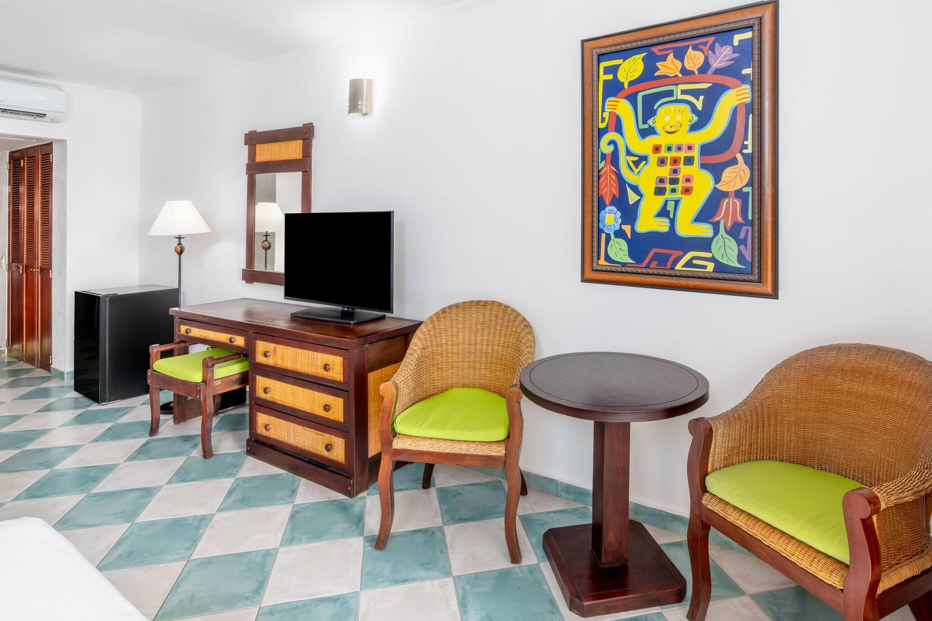 Una habitación con televisión, muebles de madera, un sillón de mimbre con cojín verde y una obra de arte colorida en la pared.