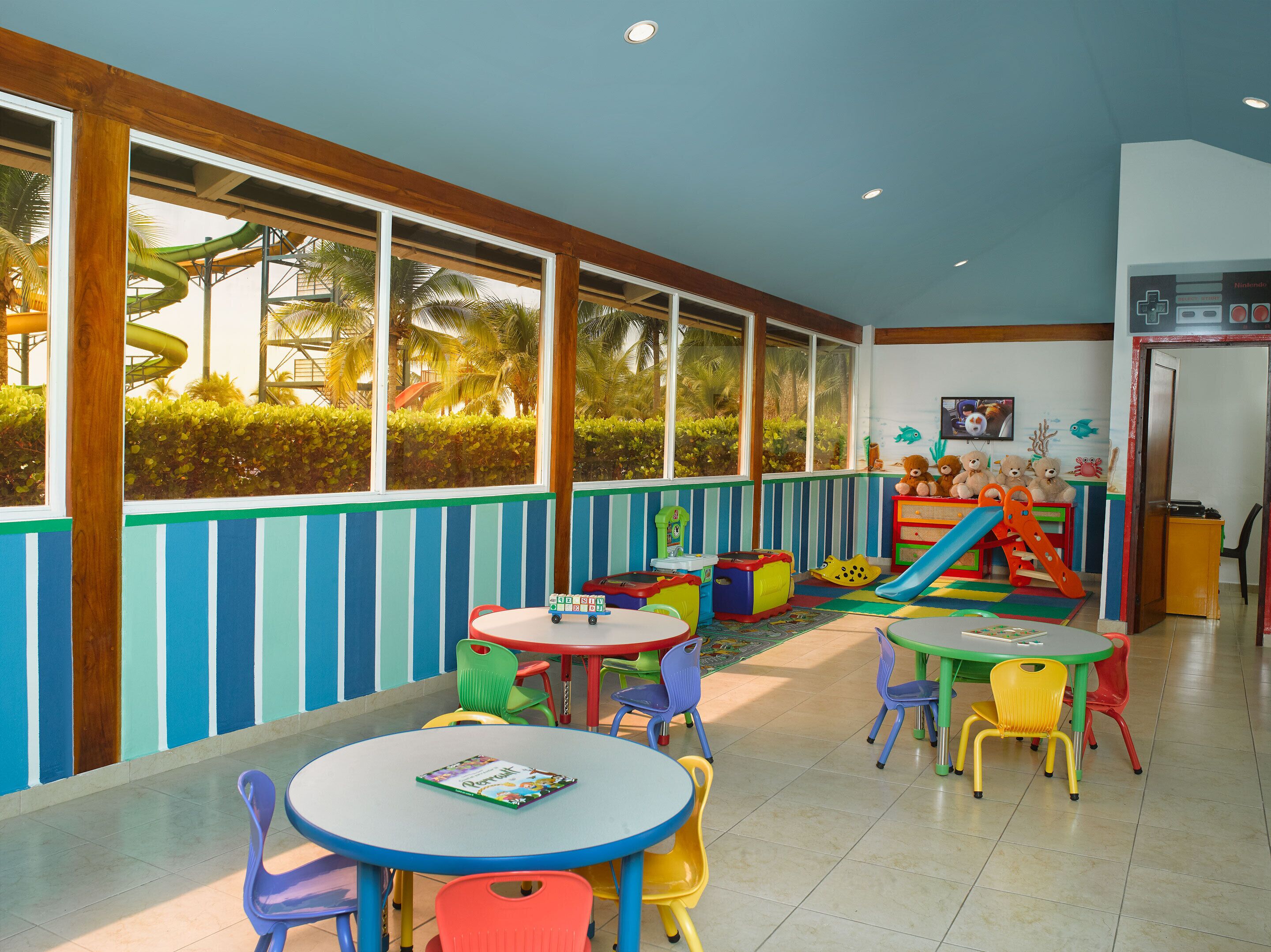Zona de juegos infantiles con mobiliario colorido y un tobogán, ventanas amplias con vistas a palmeras y el logo de Nintendo en la pared.