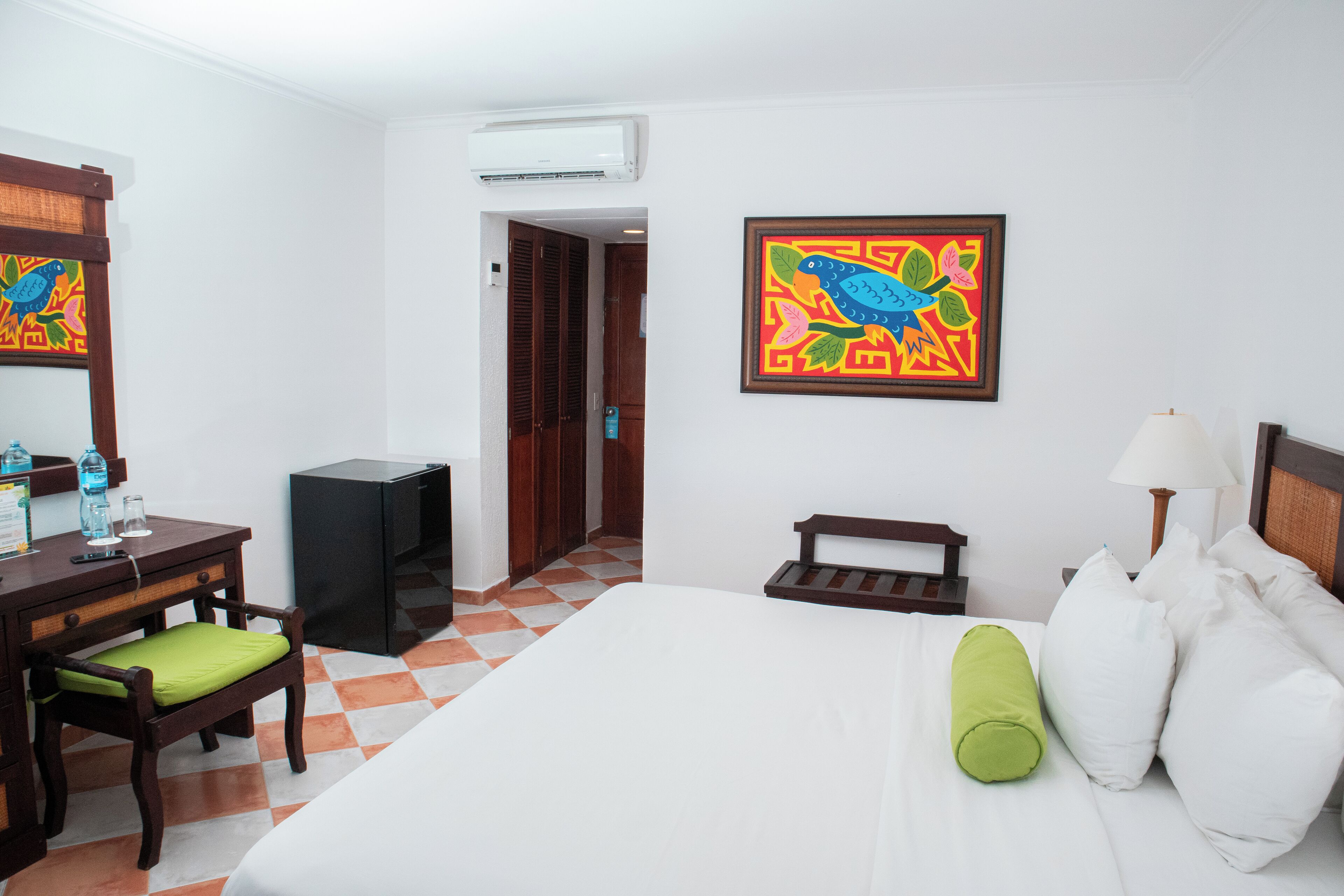 A hotel room with a bed, a desk with a chair, a small table, a lamp, and a colorful painting on the wall.