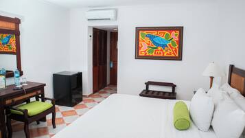 A hotel room with a bed, a desk with a chair, a small table, a lamp, and a colorful painting on the wall.