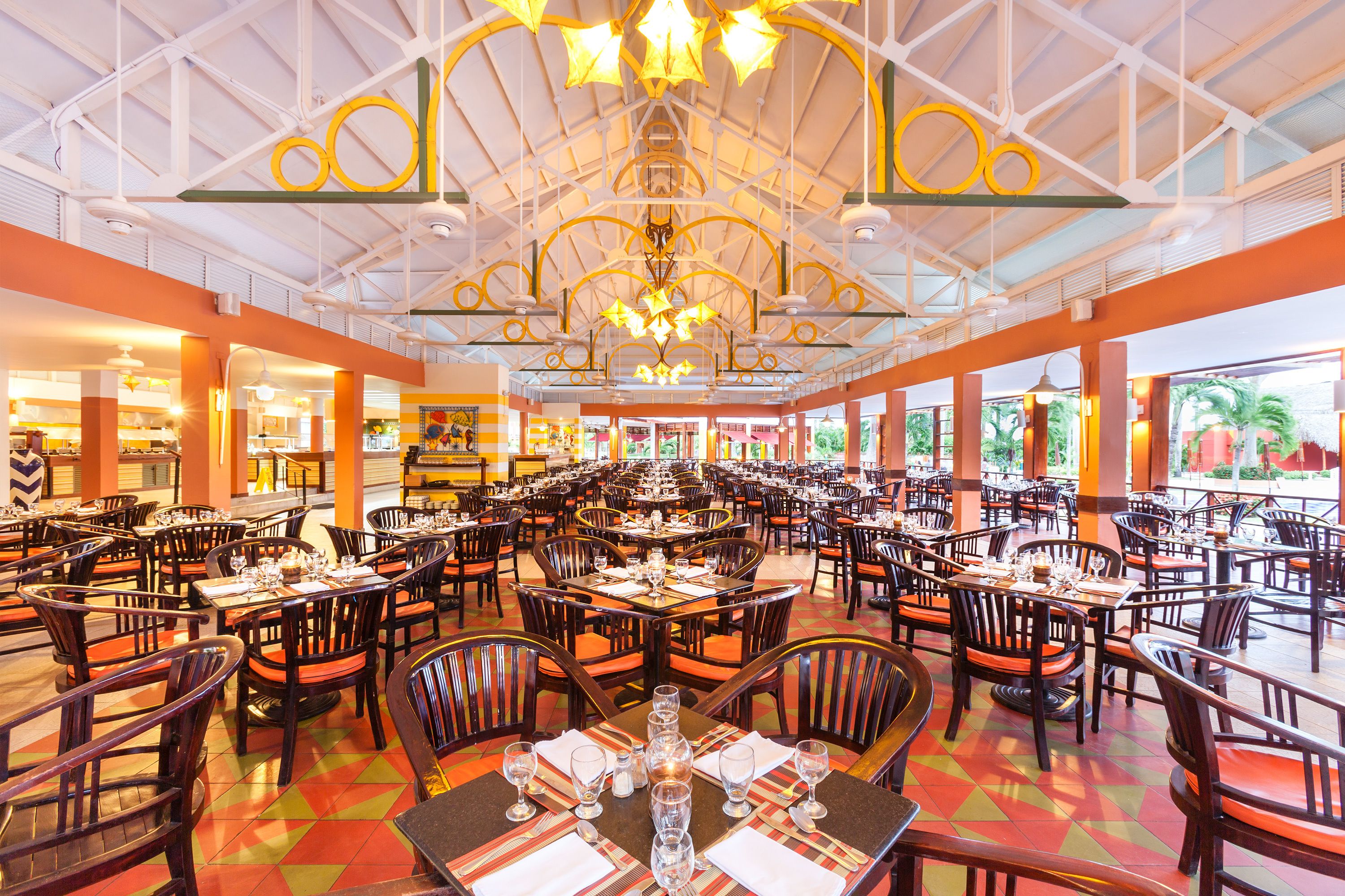 A spacious restaurant with multiple dining tables and chairs, a high ceiling with decorative lighting, and large windows offering views of the outdoors.