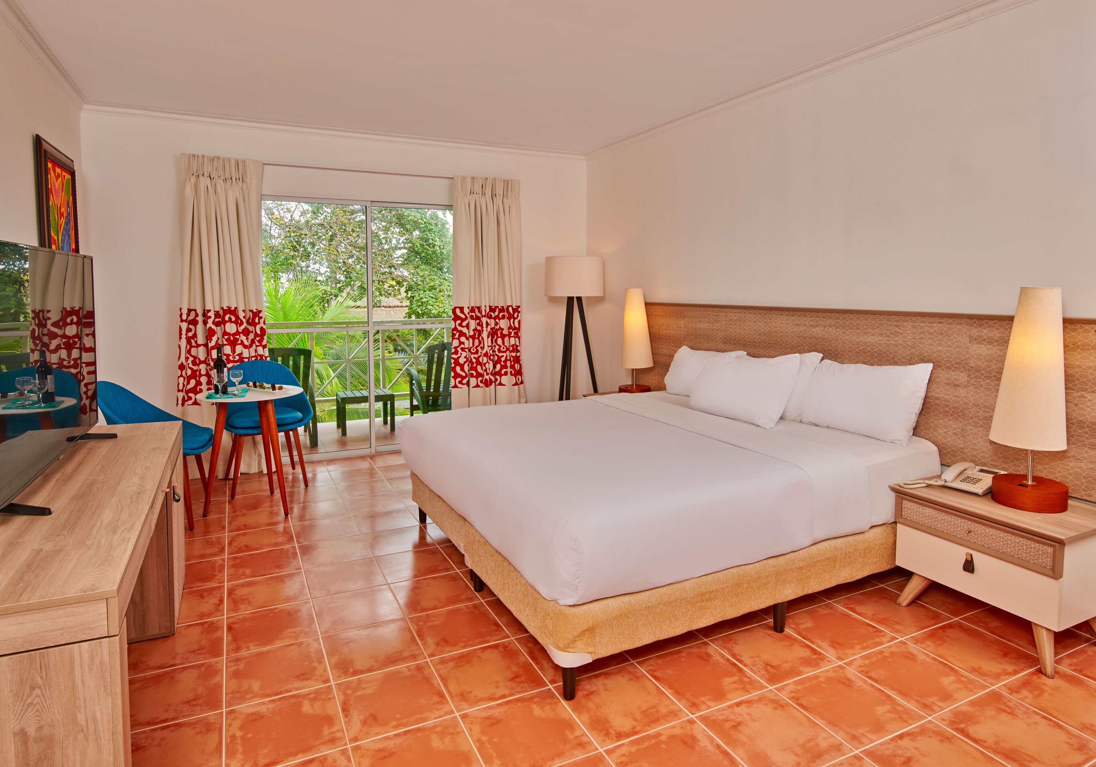 Habitación de hotel con cama, mesita de noche, lámpara, escritorio, silla y vista a follaje a través del ventanal.