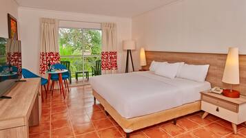 Quarto de hotel com cama, mesa de cabeceira, luminária, escrivaninha, cadeira e vista para o verde pela janela.