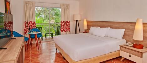 Habitación de hotel con cama, mesita de noche, lámpara, escritorio, silla y vista a follaje a través del ventanal.