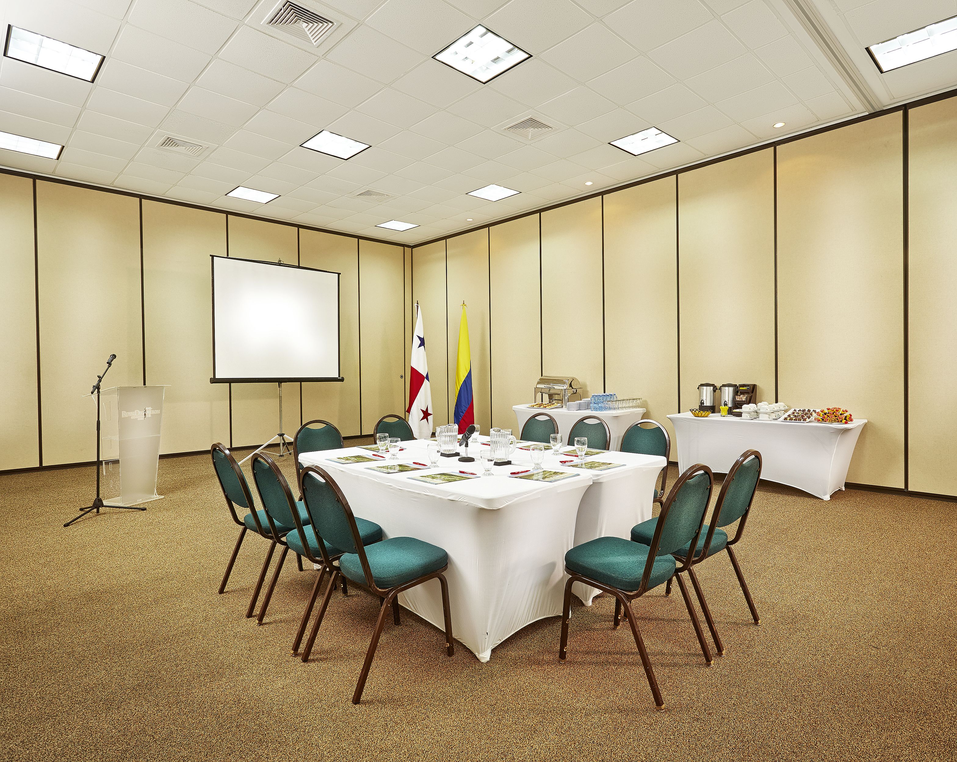A conference room with a white table, green chairs, a podium, a projector screen, and flags.