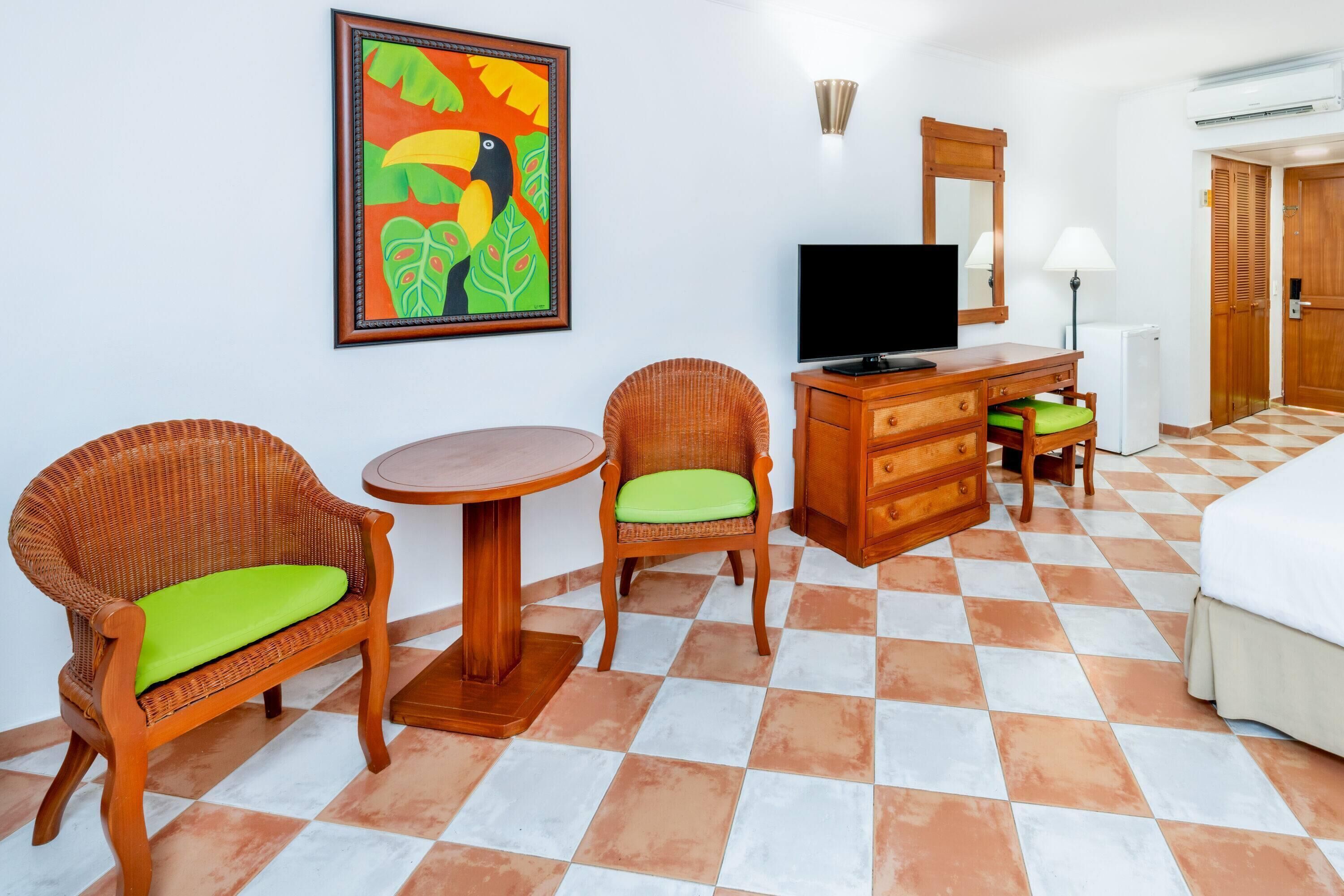 A hotel room with two wicker chairs with green cushions, a wooden dresser, a flat-screen TV, a round table, a bed with white bedding, and a tiled floor.