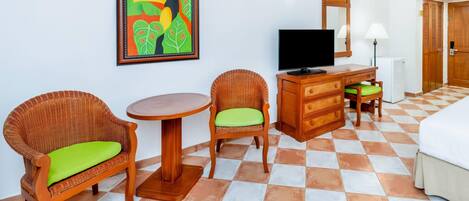 Hotelkamer met twee rieten stoelen met groene kussens, een houten dressoir, een flatscreen televisie, een ronde tafel, een bed met wit beddengoed en een betegelde vloer.