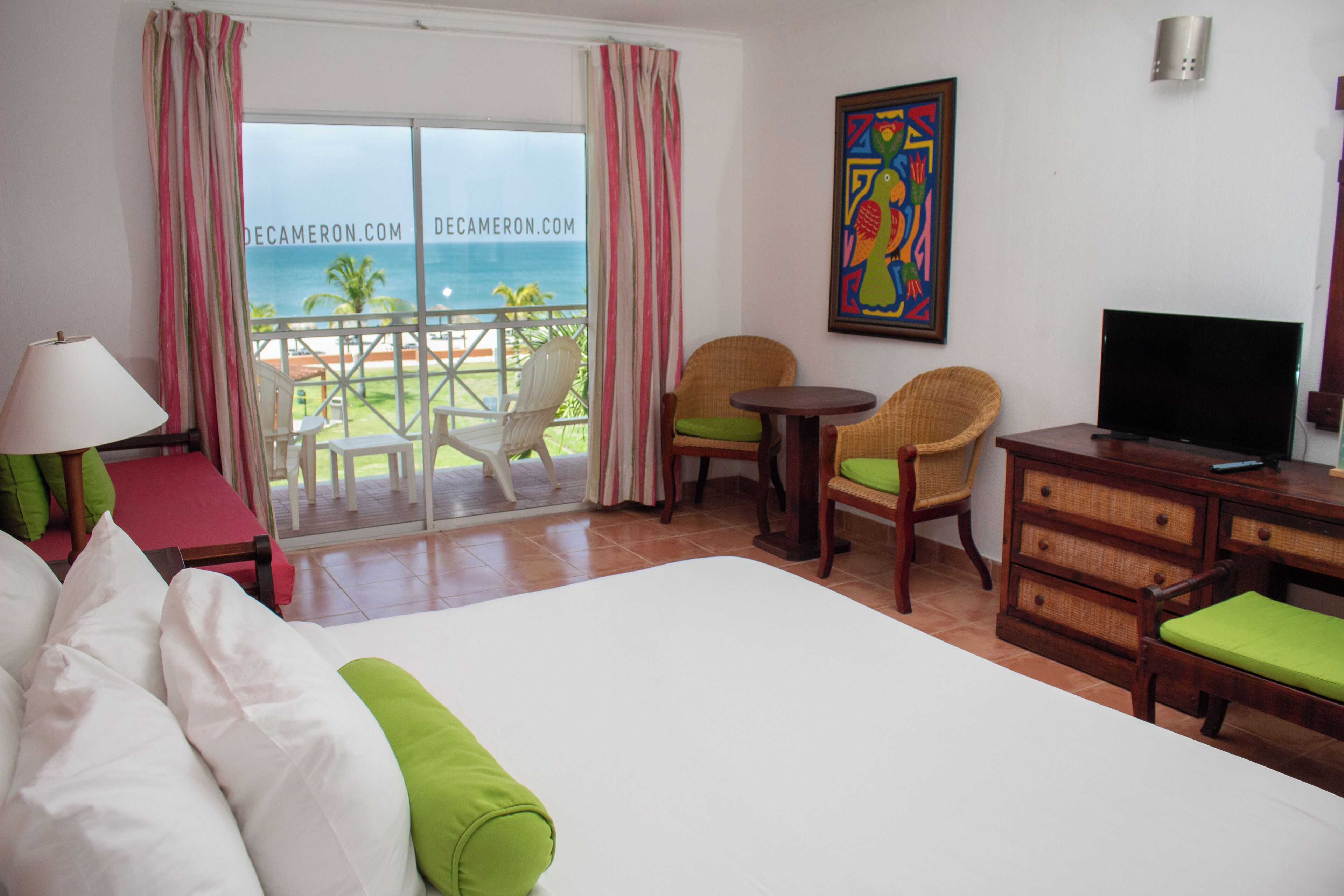 A hotel room with a bed, a TV, a desk, two chairs, a lamp, and a view of the ocean.