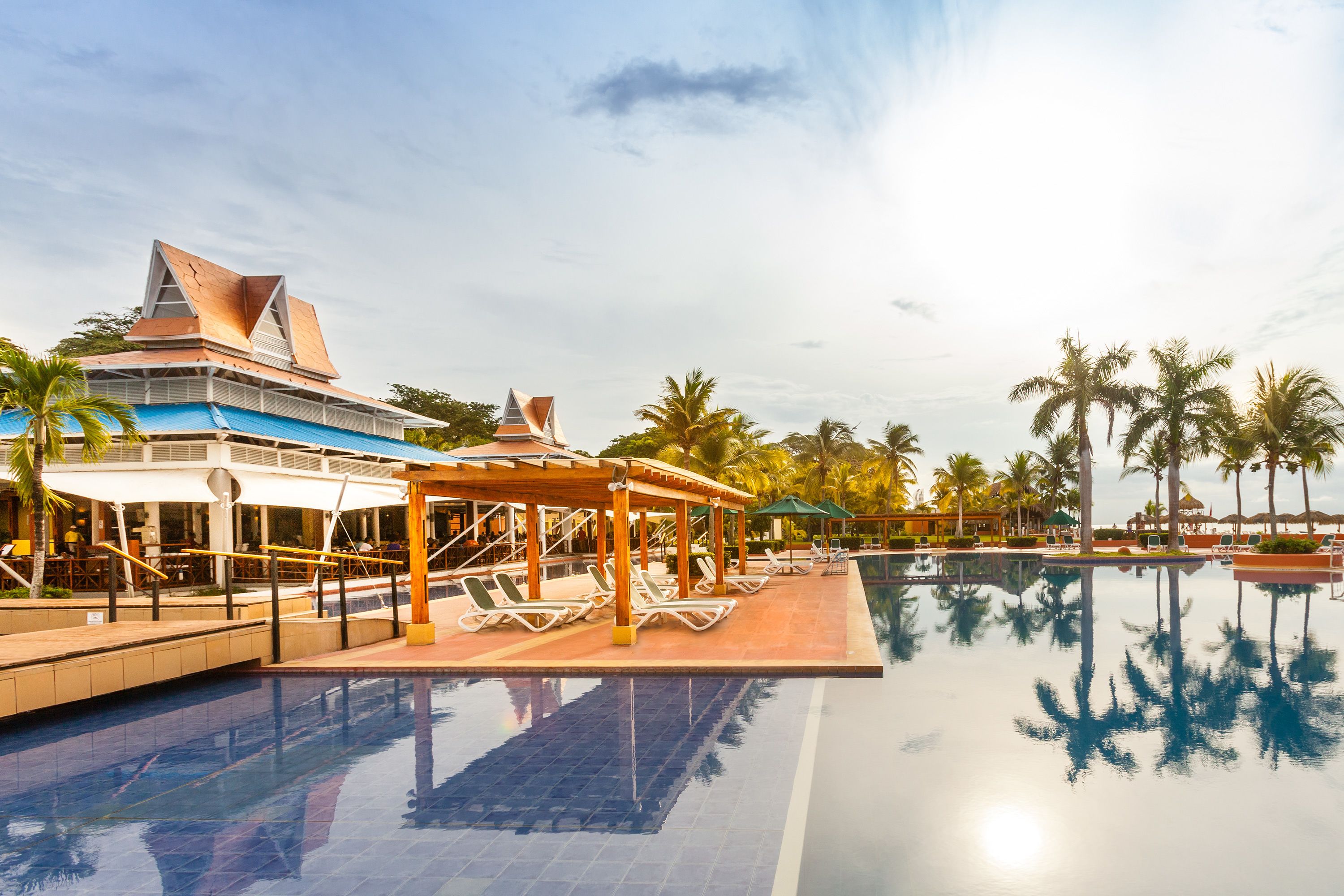Een zwembadgedeelte van een resort met ligbedden, een overdekte zithoek en palmbomen.