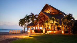 Un resort frente a la playa con una construcción de techo de paja, palmeras y una playa con sillas de descanso.