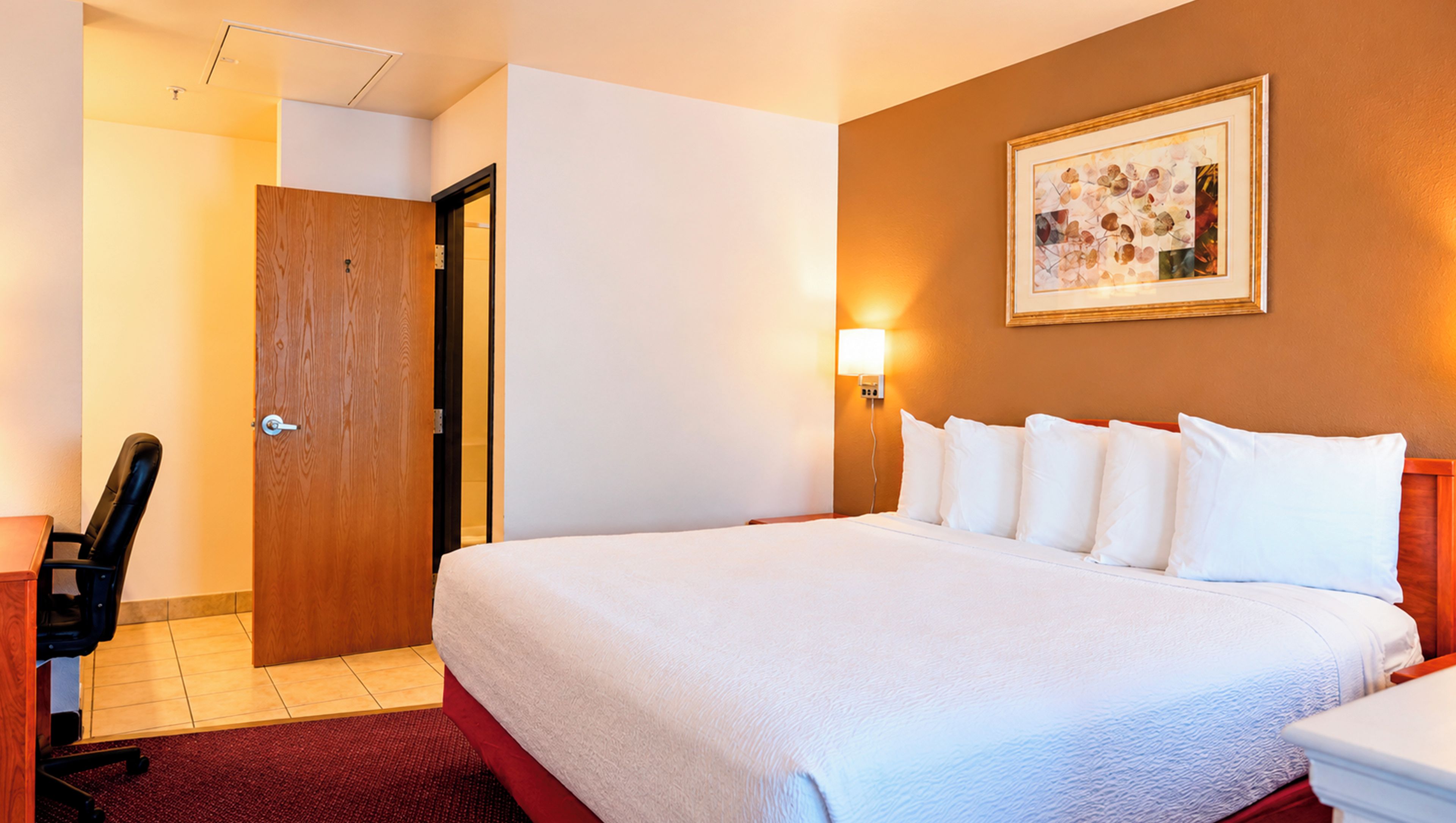 Una habitación de hotel con cama, escritorio, silla, puerta y una lámpara fijada a la pared.