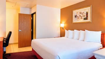 Una habitación de hotel con cama, escritorio, silla, puerta y una lámpara fijada a la pared.