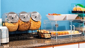Un buffet de petit-déjeuner avec des contenants étiquetés contenant des céréales et une vitrine réfrigérée où sont présentés des muffins.