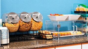 Un bufé de desayuno con recipientes etiquetados de cereales y una vitrina con muffins.
