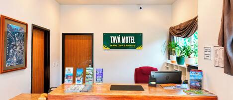 Een receptiebalie met een bordje voor Tava Motel, een rode stoel, een laptop en een plant.