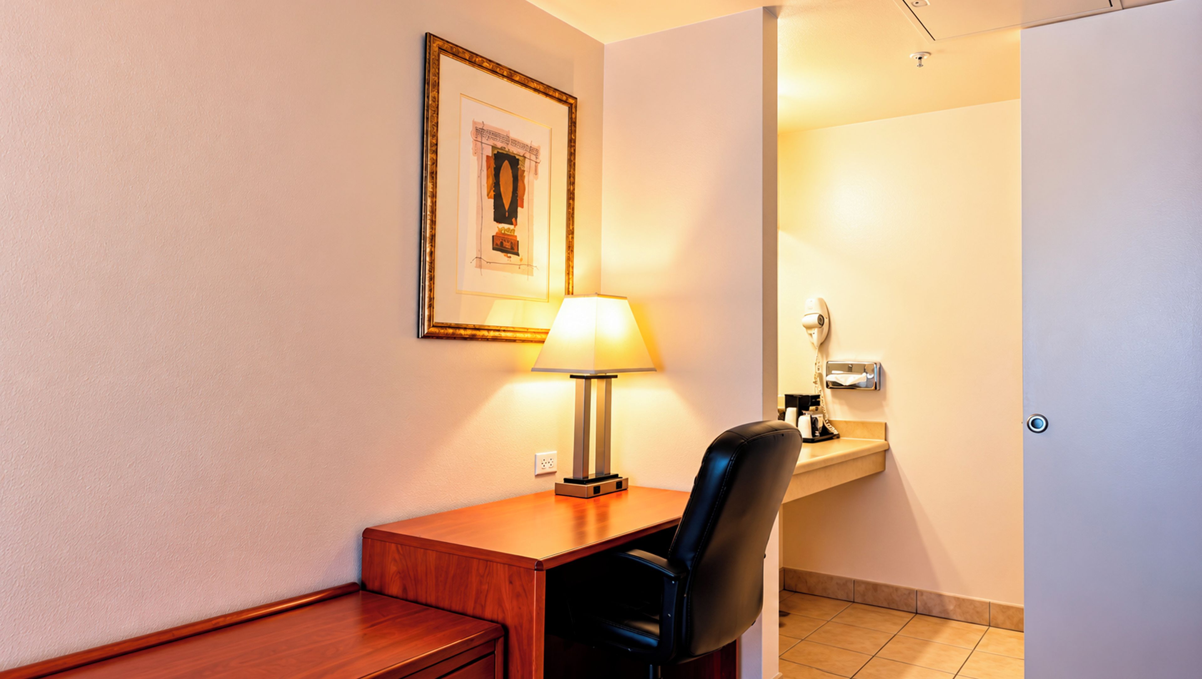 A hotel room with a wooden desk, a black leather chair, a framed picture on the wall, a bedside lamp, a small table with toiletries, and a door leading to a bathroom.