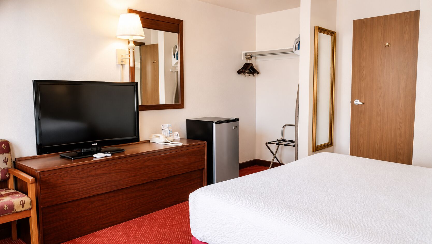 A hotel room with a bed, a TV on a wooden dresser, a desk with a lamp, a mirror, a coat rack, and a door.