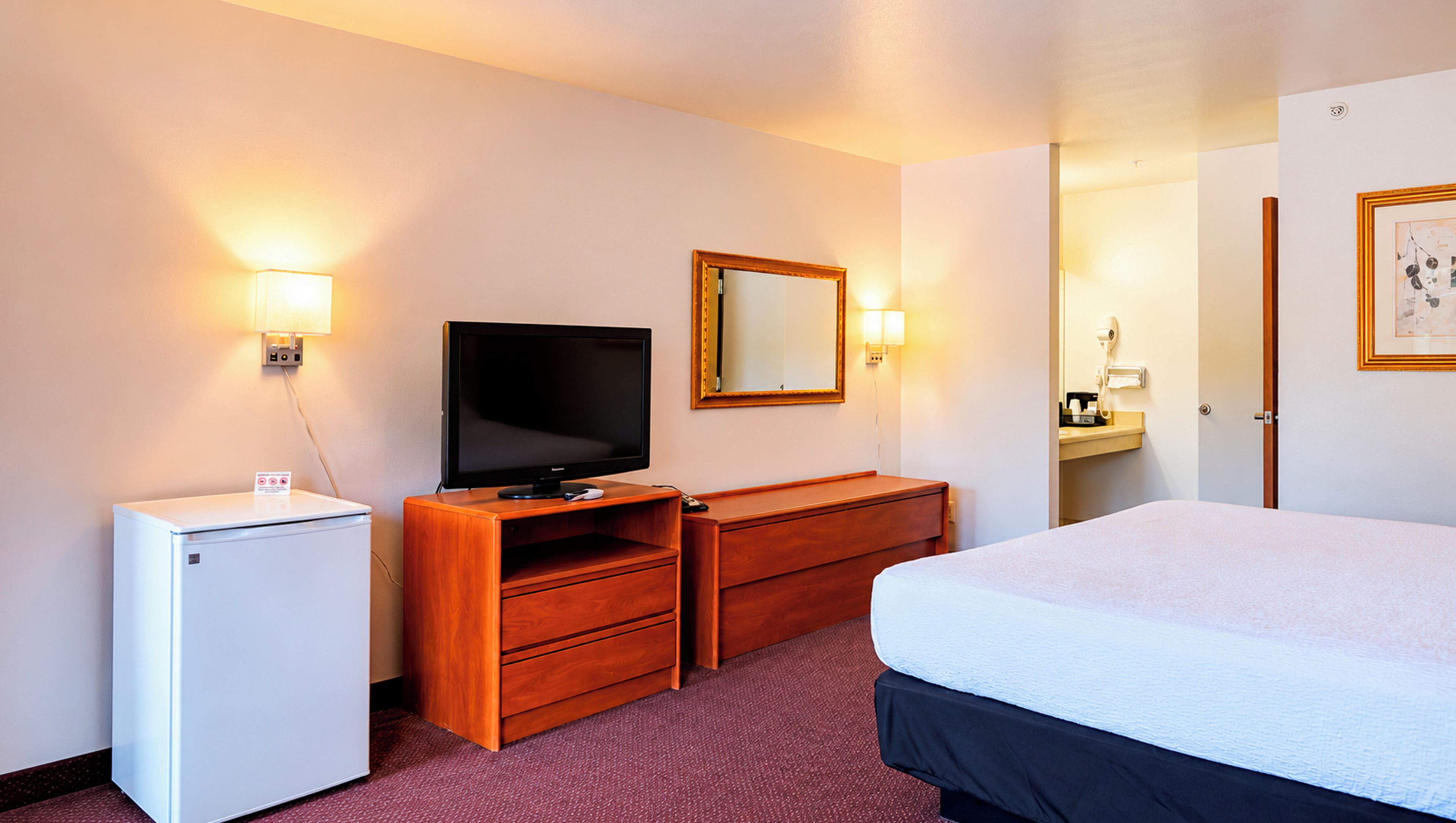 Habitación de hotel con cama, televisor, cómoda de madera, refrigerador pequeño y espejo.