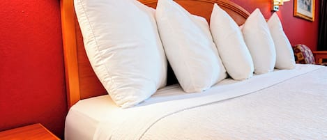 A neatly made bed with white bedding and pillows, a wooden headboard, and a red wall with a framed picture and a wall lamp.