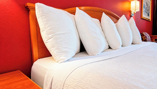 Un lit bien fait, avec une literie et des oreillers blancs, une tête de lit en bois et un mur rouge orné d’un tableau encadré et d’une lampe murale.