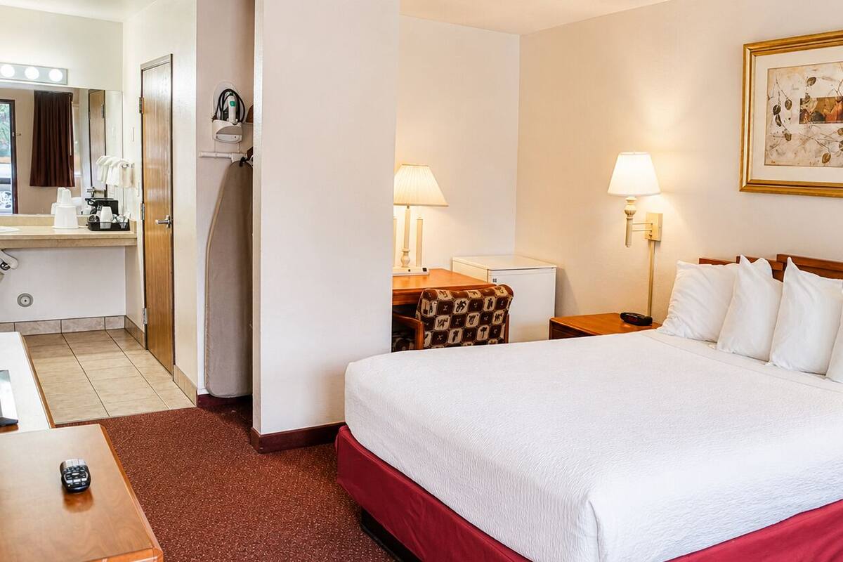 Habitació d'hotel amb llit, taules de nit, escriptori, cadira, làmpada, quadre i bany amb dutxa i mirall.