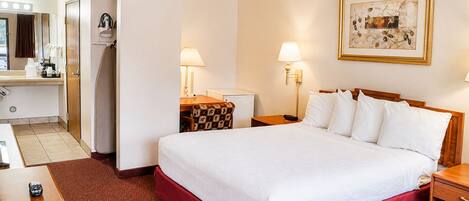 Une chambre d’hôtel comprenant un lit, des tables de chevet, un bureau, une chaise, une lampe, un tableau et une salle de bain avec douche et miroir.