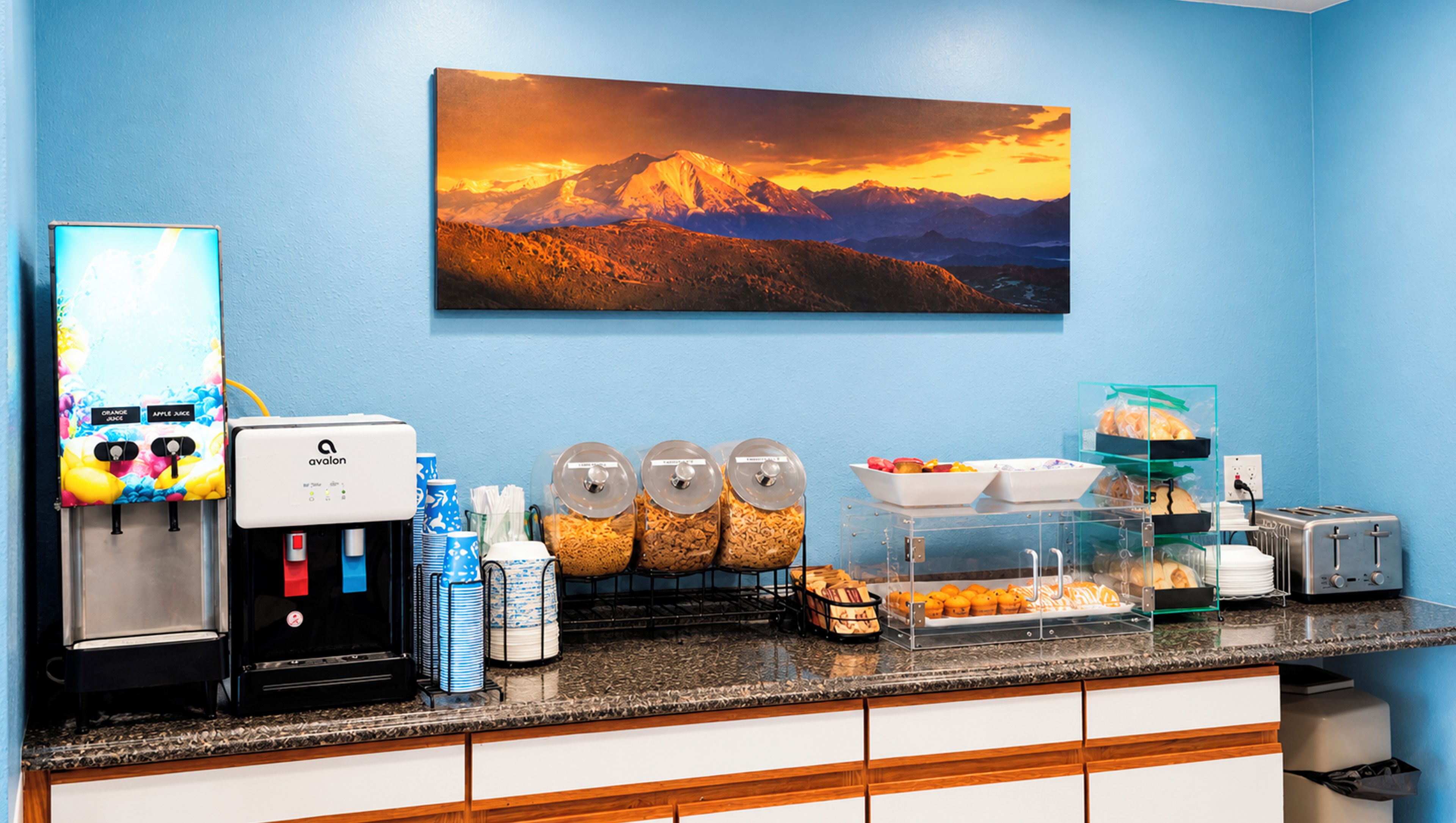 A hotel breakfast area with a coffee machine, juice dispenser, cereal dispensers, and a toaster.