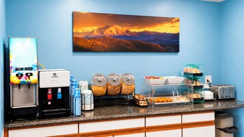 A hotel breakfast area with a coffee machine, juice dispenser, cereal dispensers, and a toaster.