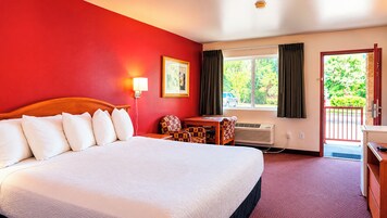 A hotel room with a large bed, a desk, a chair, a window with curtains, and a door leading to a balcony.