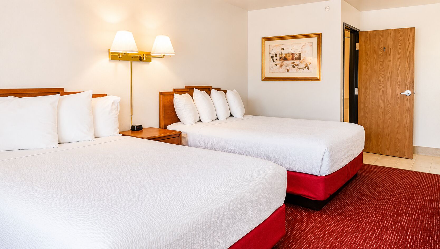Habitación de hotel con dos camas, alfombra roja, lámpara de pared y un cuadro enmarcado.